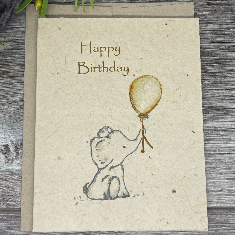 A folded plantable birthday card with hand-drawn watercolor art of an elephant holding a balloon, accompanied by the text 'Happy Birthday' on the front. Plantable card. elephant. childrens birthday.