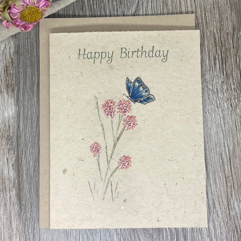 A folded plantable birthday card with hand-drawn watercolor illustrations of pink flowers and a blue butterfly on the front, labeled 'Happy Birthday'. plantable card. wildflower seed paper.