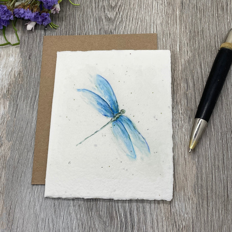 A square watercolor-style dragonfly artwork on textured white paper, placed on top of a brown kraft envelope or card, with a black-and-silver pen and small purple flowers on a gray wood surface.