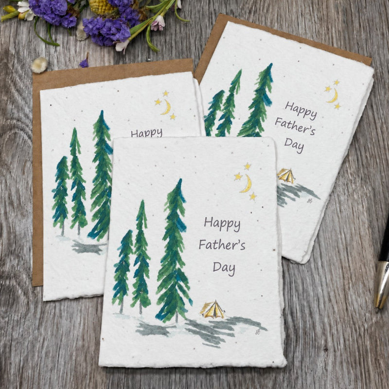 Three Father's Day greeting cards with a watercolor pine tree and campfire design, arranged on a rustic wooden surface with a black and gold pen and small flowers.