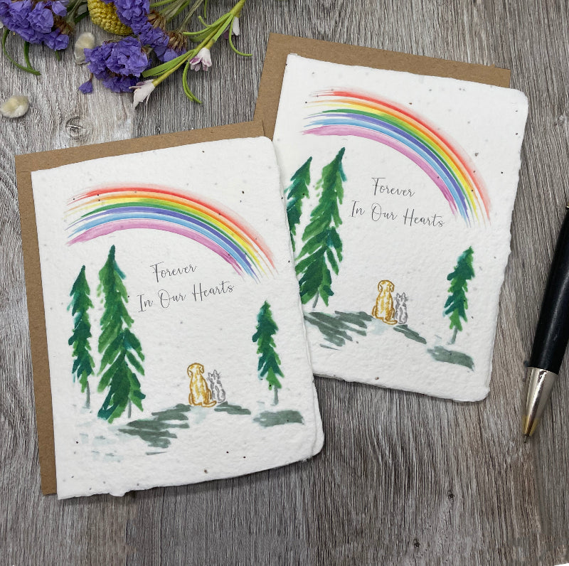 Two square handmade paper cards with brown kraft envelopes, featuring a watercolor rainbow, pine trees, and a small dog, with the text 'Forever In Our Hearts', arranged on a gray wood surface with purple flowers and a black pen nearby.