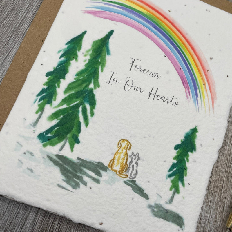 A square white greeting card with a watercolor rainbow, green pine trees, and the words 'Forever In Our Hearts,' placed on a brown kraft envelope over a gray wood surface.